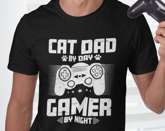 May include: Black t-shirt with a white graphic design of a video game controller and the text "Cat Dad By Day Gamer By Night".