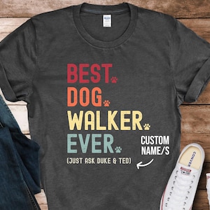 May include: Dark grey t-shirt with a vintage-style graphic that reads "Best Dog Walker Ever (Just Ask Duke & Ted)" with a paw print after each word. The text "Custom Name/S" is written below the graphic with an arrow pointing to it.