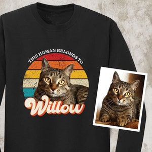 May include: Black long-sleeved shirt featuring a retro-style graphic of a cat with the text "This human belongs to Willow". The graphic includes a sunset with stripes of orange, yellow, blue, and red. A smaller image of the cat is in the corner of the shirt.