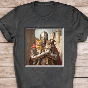 May include: A charcoal gray t-shirt featuring a graphic print of a medieval knight in full metal armor and a red cape, holding four small cats in their arms. The knight stands before a stone castle with towers under a cloudy sky.