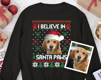 May include: A black long-sleeve sweatshirt featuring a graphic of a golden dog wearing a Santa hat. The text reads I BELIEVE IN SANTA PAWS, surrounded by red and green holiday patterns including paw prints, snowflakes, and Christmas trees.