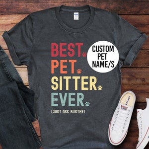 May include: A dark grey t-shirt with the text "BEST PET SITTER EVER (JUST ASK BUSTER)" in a vintage style font. The text is in red, yellow, green, and blue. There is a paw print after each word. There is a white circle with the text "CUSTOM PET NAME/S" in black text.