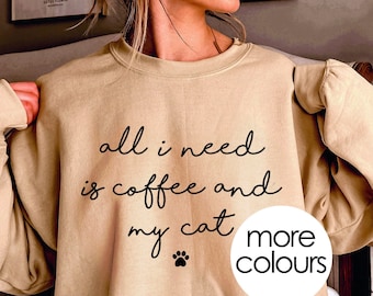 May include: A beige sweatshirt with the text "all i need is coffee and my cat" in black. A paw print is below the text. The text "more colours" is in a white circle with a black outline.