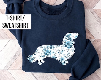 May include: A navy blue long-sleeve sweatshirt featuring a white silhouette of a long-haired dog filled with a blue floral pattern. The text T-SHIRT/SWEATSHIRT is printed in black on a white rectangular background in the upper left corner.