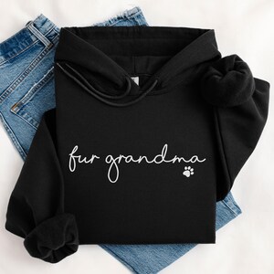 Puede incluir: Sudadera con capucha negra con la frase "fur grandma" en blanco, acompa&ntilde;ada de una huella de pata. La sudadera se muestra sobre un fondo claro con vaqueros azules.