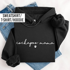 May include: A black hooded sweatshirt featuring the white text "cockapoo mama" written in a cursive script font, accompanied by a small white paw print graphic centered below the text. The hoodie includes a drawstring hood and long sleeves.