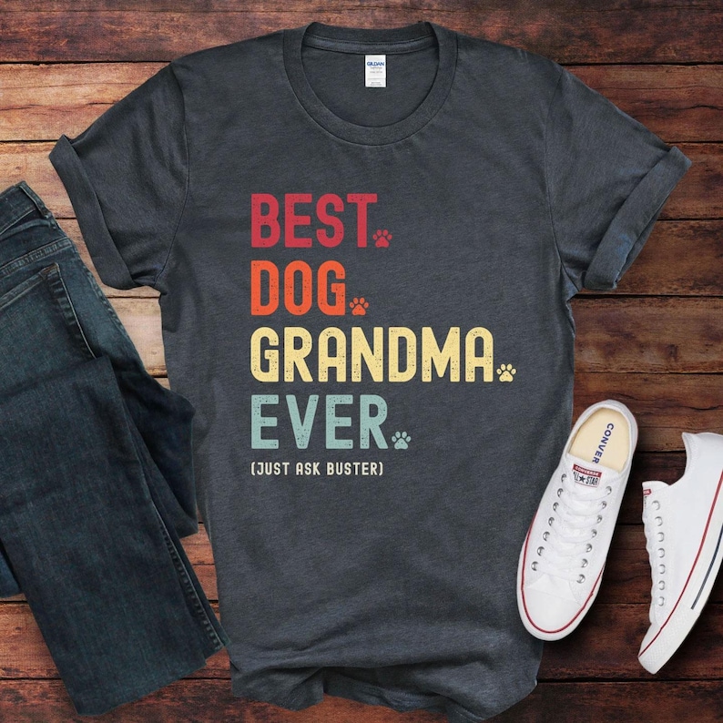 Puede incluir: Camiseta gris oscuro con el texto "BEST DOG GRANDMA EVER" en rojo, naranja, amarillo y azul. La camiseta incluye huellas de patas y la frase "(JUST ASK BUSTER)". Un par de vaqueros azules y zapatillas blancas est&aacute;n junto a la camiseta.