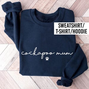 May include: A navy blue long-sleeve sweatshirt featuring the white text "cockapoo mum" in a cursive script font, accompanied by a small white paw print graphic. The upper right corner displays the text "SWEATSHIRT/T-SHIRT/HOODIE".