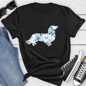 May include: A black short-sleeved t-shirt featuring a graphic silhouette of a long-haired dog filled with a blue and white floral pattern. The shirt is displayed alongside a pair of blue denim jeans, black sunglasses, and black and white sneakers.