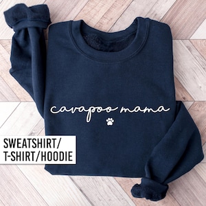 May include: Navy blue sweatshirt with the words "cavapoo mama" in white script and a small paw print. The bottom of the sweatshirt is cuffed. The image also includes the text "SWEATSHIRT/ T-SHIRT/ HOODIE".