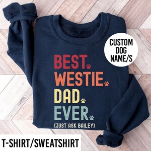 May include: Navy blue sweatshirt with the text "BEST WESTIE DAD EVER" in red, orange, yellow, and green. The text includes paw prints and the phrase "(JUST ASK BAILEY)". A white circle reads "CUSTOM DOG NAME/S". The bottom of the image reads "T-SHIRT/SWEATSHIRT".