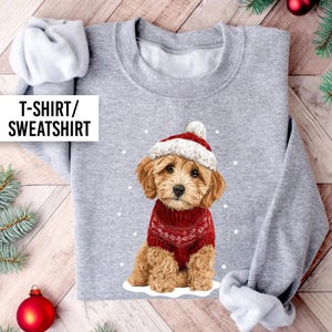 May include: A grey sweatshirt with a festive illustration of a puppy wearing a red sweater and a matching hat with a white pom-pom. The text "T-SHIRT/ SWEATSHIRT" is in a white box on the left. Snowflakes are scattered around the puppy.