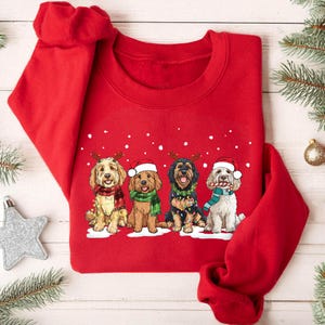 May include: A bright red crewneck sweatshirt featuring a graphic of four dogs wearing holiday accessories. The dogs are adorned with Santa hats, scarves, and string lights, set against a background of white falling snow on the fabric.