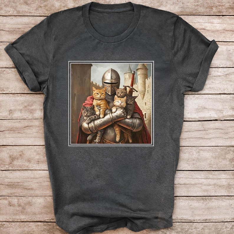 May include: A charcoal gray t-shirt featuring a graphic print of a medieval knight in full metal armor and a red cape, holding four small cats in their arms. The knight stands before a stone castle with towers under a cloudy sky.