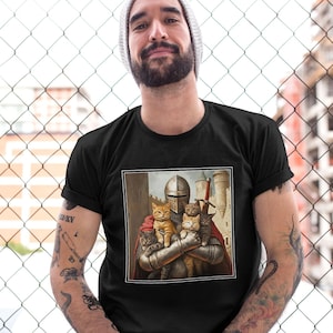 May include: A black short-sleeved t-shirt featuring a square graphic print of a medieval knight in full metal armor holding four small cats. The knight wears a red cape and stands in front of a stone castle structure with a sword visible.