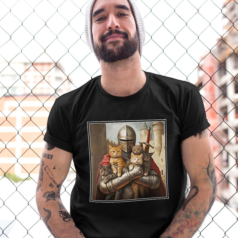 May include: A black short-sleeved t-shirt featuring a square graphic print of a medieval knight in full metal armor holding four small cats. The knight wears a red cape and stands in front of a stone castle structure with a sword visible.