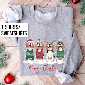 May include: A grey sweatshirt with a festive design. It features four cartoon beagles dressed in Christmas attire, with the words "Merry Christmas" in red. The text "T-SHIRTS/ SWEATSHIRTS" is in a white box in the upper left corner.