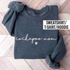May include: A dark heather grey long-sleeve sweatshirt with the white text "cockapoo mom" written in a cursive script font, accompanied by a small white paw print graphic. The text "SWEATSHIRT/T-SHIRT/HOODIE" is printed in a white box.