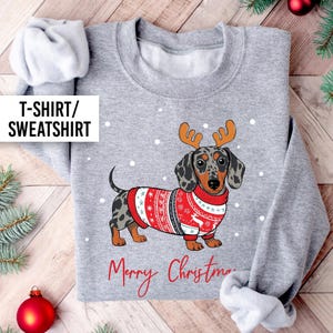 May include: A grey sweatshirt with a dachshund wearing a festive red and white Christmas sweater and antlers. The words "Merry Christmas" are written in red script. The text "T-SHIRT/ SWEATSHIRT" is in a white box.