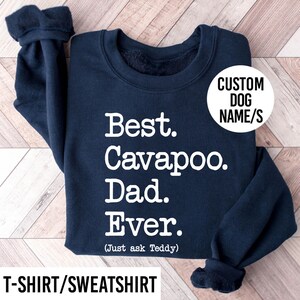 May include: A navy blue long-sleeve sweatshirt featuring white text that reads Best. Cavapoo. Dad. Ever. (Just ask Teddy). A circular white graphic overlay in the upper right corner contains the black text CUSTOM DOG NAME/S.