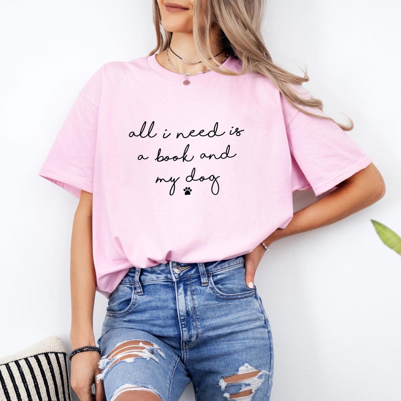 May include: A light pink short-sleeved t-shirt featuring the black printed text "all i need is a book and my dog" with a small paw print icon centered on the chest. The shirt has a crew neckline and a relaxed fit, paired with blue denim jeans.