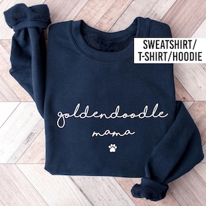 May include: A navy blue long-sleeve sweatshirt featuring the white text "goldendoodle mama" in a cursive script font, accompanied by a small white paw print graphic below the text. The upper right corner includes the text "SWEATSHIRT/T-SHIRT/HOODIE".
