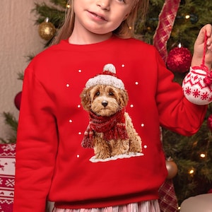May include: A bright red long-sleeve sweatshirt featuring a graphic print of a fluffy brown puppy wearing a white-trimmed red Santa hat and a matching red scarf, surrounded by small white dots resembling falling snow against the red fabric.