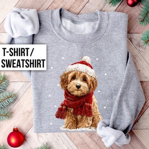 May include: A gray sweatshirt featuring a cute puppy wearing a Santa hat and a red scarf. The text "T-SHIRT / SWEATSHIRT" is displayed on the left side of the image. The sweatshirt is decorated with white snowflakes.