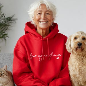 Puede incluir: Una sudadera con capucha roja con la frase "fur grandma" en escritura blanca y una huella de pata. Una mujer con cabello blanco sonr&iacute;e, flanqueada por un golden doodle y un gato atigrado.
