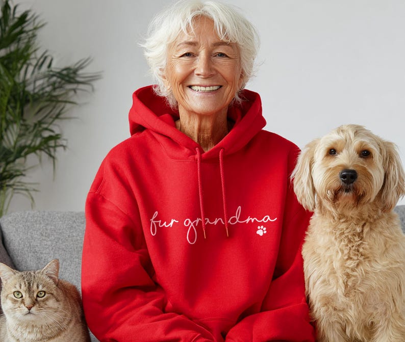 Puede incluir: Una sudadera con capucha roja con la frase "fur grandma" en escritura blanca y una huella de pata. Una mujer con cabello blanco sonr&iacute;e, flanqueada por un golden doodle y un gato atigrado.