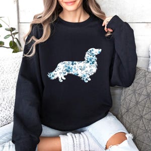May include: A black long-sleeve crewneck sweatshirt featuring a graphic of a dog silhouette filled with a pattern of blue and white flowers. The dog is depicted in profile, facing right, with a detailed floral print covering its entire body.