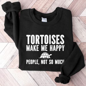 May include: Black sweatshirt with white text: "TORTOISES MAKE ME HAPPY PEOPLE, NOT SO MUCH" and a tortoise graphic. The sweatshirt is folded on a light wood grain surface.