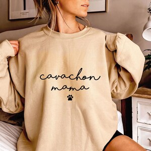 May include: A beige crewneck sweatshirt featuring the black text "cavachon mama" written in a cursive script font, positioned above a small black paw print graphic centered on the chest of the garment, which has long sleeves and a ribbed neckline.