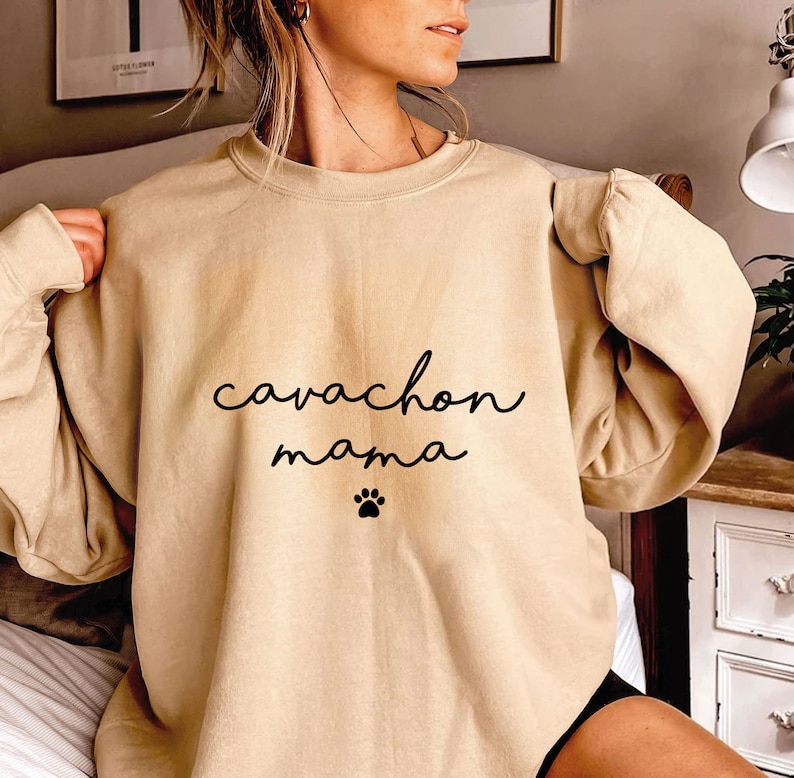 May include: A beige crewneck sweatshirt featuring the black text "cavachon mama" written in a cursive script font, positioned above a small black paw print graphic centered on the chest of the garment, which has long sleeves and a ribbed neckline.