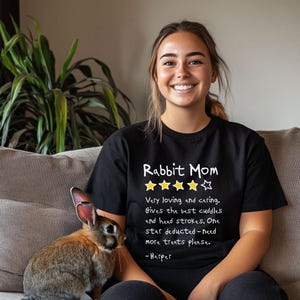 May include: A black short-sleeved t-shirt featuring the white text Rabbit Mom, four yellow stars, one white star, and the text: Very loving and caring. Gives the best cuddles and head strokes. One star deducted-need more treats please. -Harper.