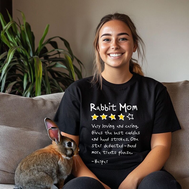 May include: A black short-sleeved t-shirt featuring the white text Rabbit Mom, four yellow stars, one white star, and the text: Very loving and caring. Gives the best cuddles and head strokes. One star deducted-need more treats please. -Harper.