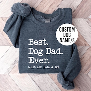 May include: A dark grey sweatshirt with the text "Best. Dog Dad. Ever." in white. The text "(Just ask Lola & Bo)" is below. A white circle with the text "CUSTOM DOG NAME/S" is on the right.