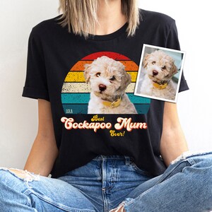 Peut inclure: Un t-shirt noir à manches courtes avec un graphique représentant un chiot clair portant un collier jaune dans un cercle de coucher de soleil rétro. Le texte indique Lola, Best Cockapoo Mum Ever! Une petite photo rectangulaire est visible.