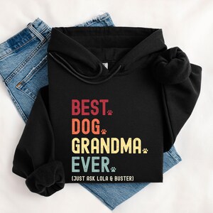 Puede incluir: Sudadera con capucha negra con el texto "BEST DOG GRANDMA EVER" en rojo, naranja, amarillo y turquesa. El dise&ntilde;o incluye huellas de patas y la frase "(JUST ASK LOLA & BUSTER)". La sudadera se muestra sobre unos vaqueros azules.