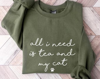 May include: An olive green crewneck sweatshirt featuring the white text all i need is tea and my cat with a small paw print icon below the lettering. The garment has long sleeves with ribbed cuffs and a ribbed neckline, presented in a flat lay.