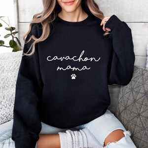 May include: A black long-sleeve crewneck sweatshirt featuring the white text "cavachon mama" in a cursive script font, positioned above a small white paw print graphic centered on the chest area of the garment.