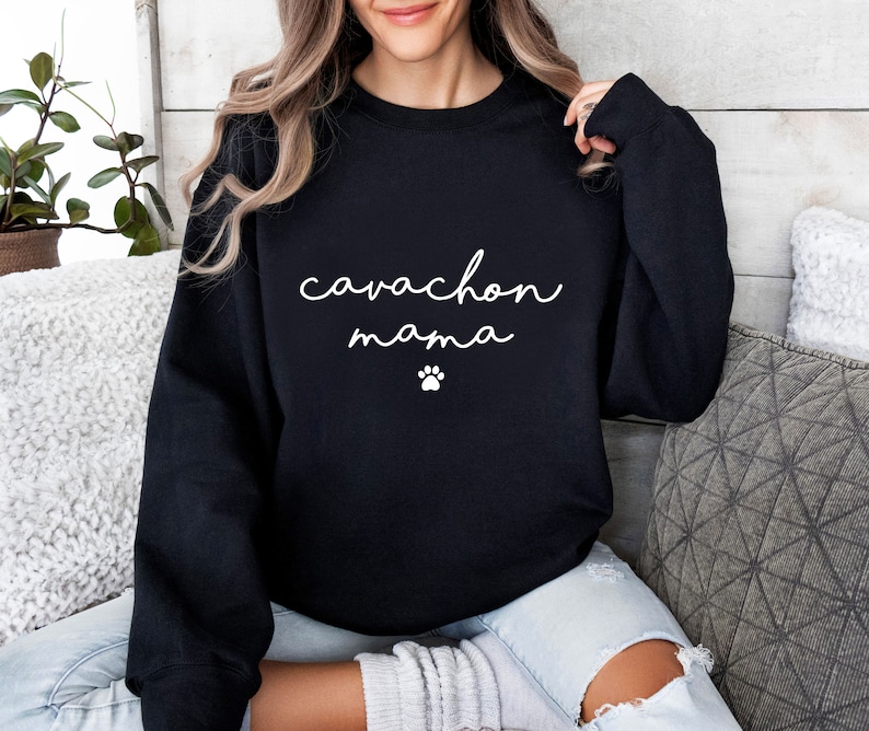 May include: A black long-sleeve crewneck sweatshirt featuring the white text "cavachon mama" in a cursive script font, positioned above a small white paw print graphic centered on the chest area of the garment.