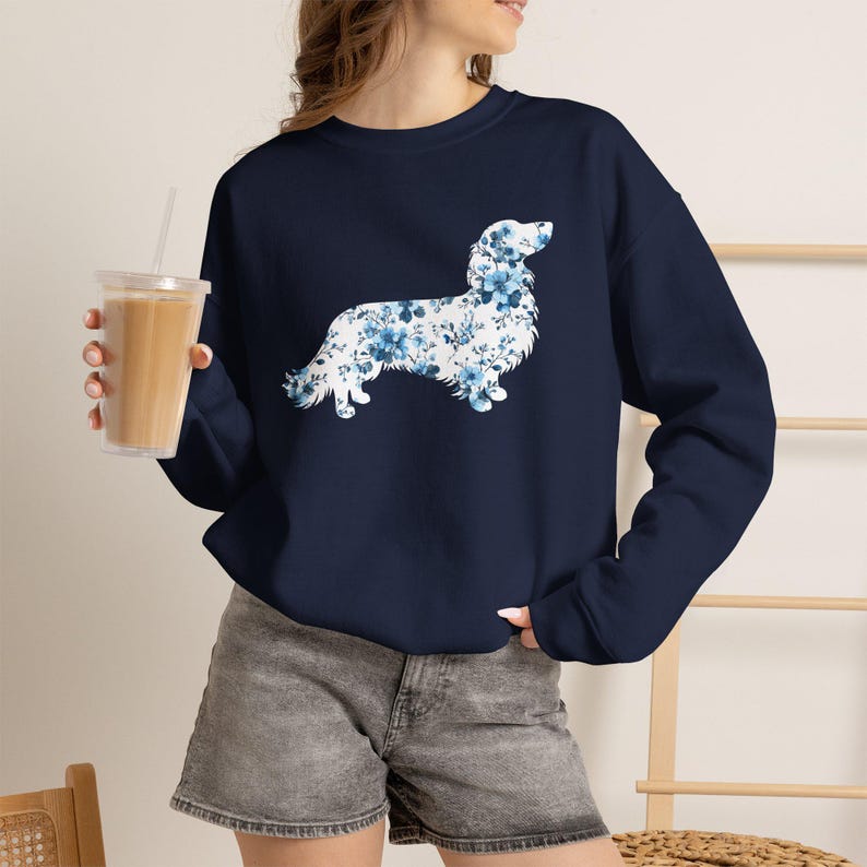 May include: A navy blue long-sleeve sweatshirt featuring a graphic of a dog silhouette filled with a blue and white floral pattern. The sweatshirt has a crew neckline and long sleeves, providing a casual look for everyday wear.