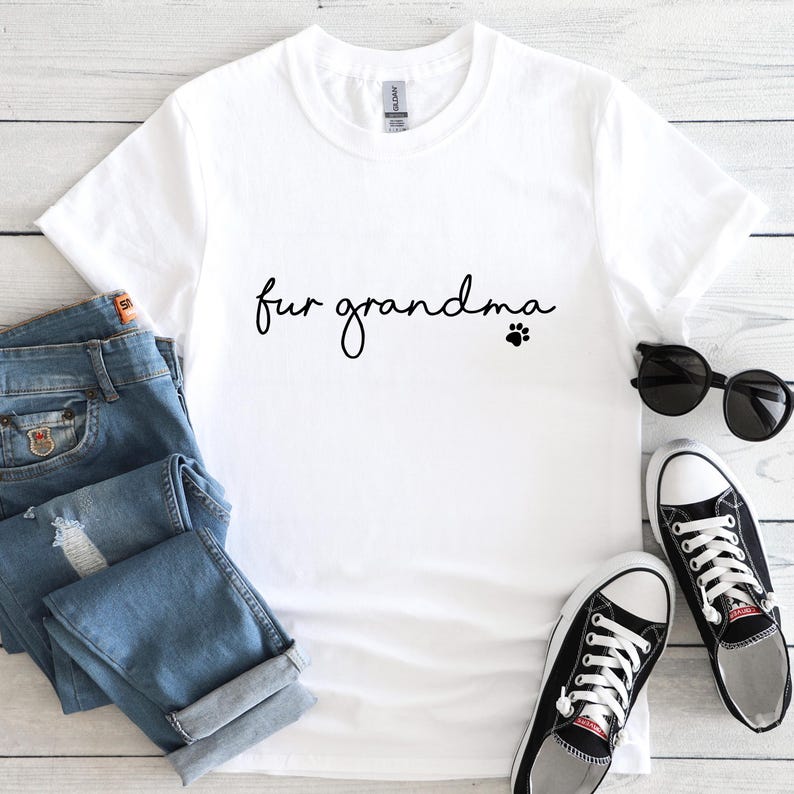 Puede incluir: Camiseta blanca con el texto "fur grandma" en fuente cursiva y una huella de pata. La camiseta se combina con vaqueros azules, gafas de sol negras y zapatillas deportivas blancas y negras.