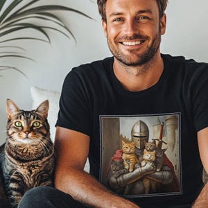 May include: A black short-sleeved t-shirt featuring a square graphic print of a medieval knight in silver armor holding four small cats in their arms. The knight wears a red cape and stands before a stone castle tower with a sword visible behind them.