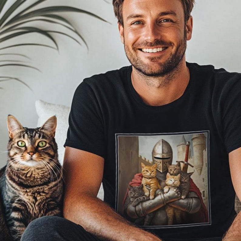 May include: A black short-sleeved t-shirt featuring a square graphic print of a medieval knight in silver armor holding four small cats in their arms. The knight wears a red cape and stands before a stone castle tower with a sword visible behind them.