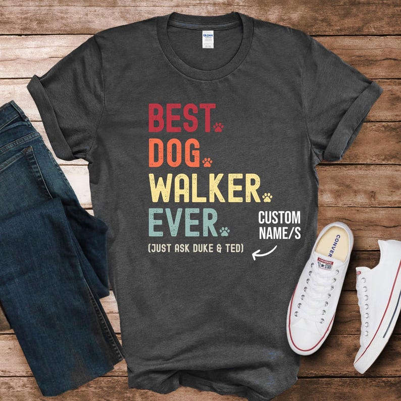 May include: A dark gray short-sleeve t-shirt featuring the text BEST DOG WALKER EVER in red, orange, yellow, and teal, with small paw prints. Below, it reads (JUST ASK DUKE & TED) and CUSTOM NAME/S with an arrow pointing to the right side.