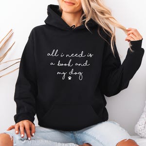 May include: A black hooded sweatshirt featuring the white text "all i need is a book and my dog" with a small paw print icon centered on the chest. The garment includes a front kangaroo pocket, long sleeves, and a drawstring hood for coverage.