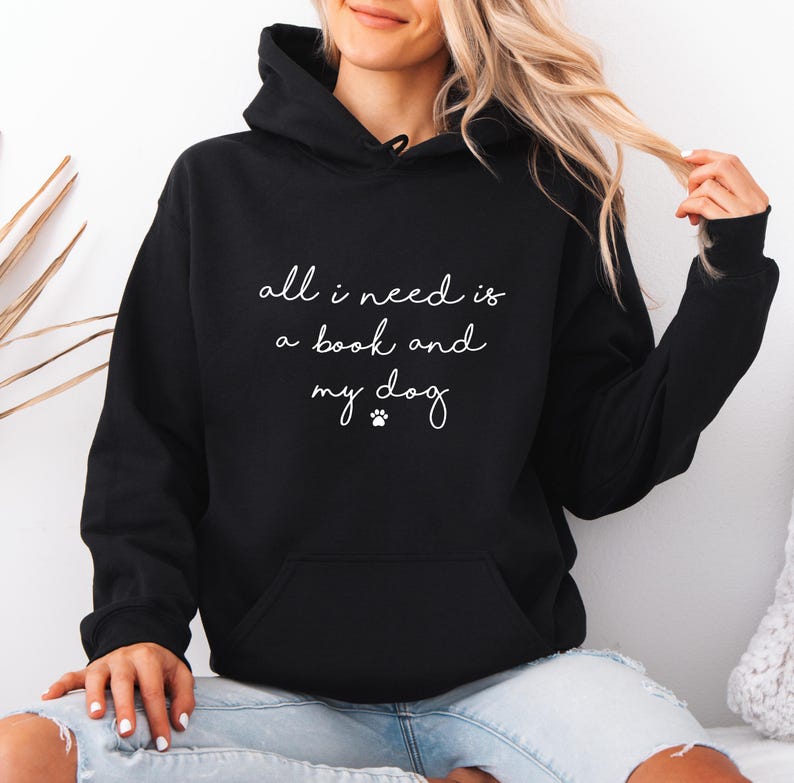 May include: A black hooded sweatshirt featuring the white text "all i need is a book and my dog" with a small paw print icon centered on the chest. The garment includes a front kangaroo pocket, long sleeves, and a drawstring hood for coverage.