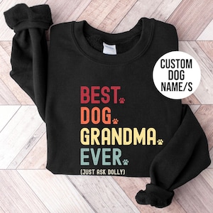 Puede incluir: Sudadera negra con el texto "BEST DOG GRANDMA EVER" en rojo, naranja, amarillo y azul. El texto incluye huellas de patas y la frase "(JUST ASK DOLLY)". Un c&iacute;rculo blanco dice "CUSTOM DOG NAME/S".